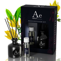 ARWA Essential Ylang-Ylang Reed Diffuser Set - black ceramic diffuser pot with reed sticks, fragrance oil bottle and refill bottle with packaging box, yellow ylang-ylang flowers in background